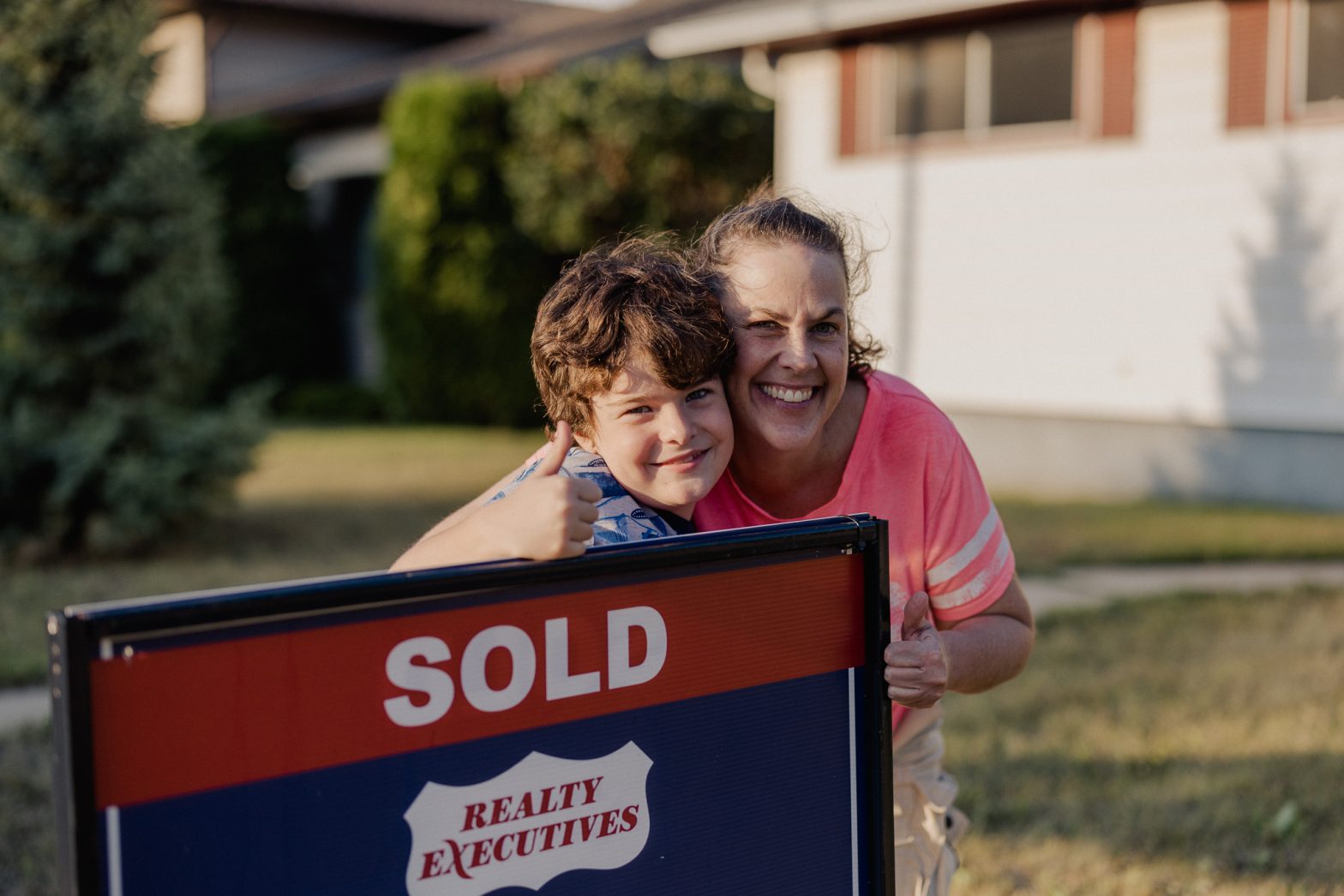 A joyful single mother and her young son standing proudly next to a 'Sold' sign, celebrating their victory of selling their home over the asking price in just four days.